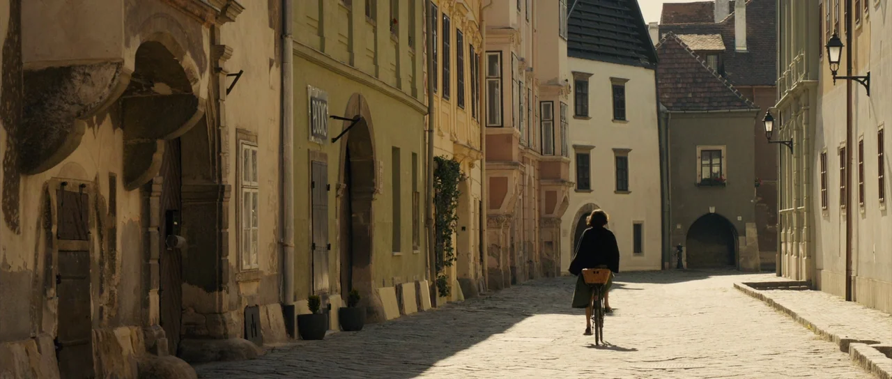 The Duke of Burgundy (2014) movie still: wide — A narrow European street is lined with historic stone buildings and a brick archway. A woman rides a…