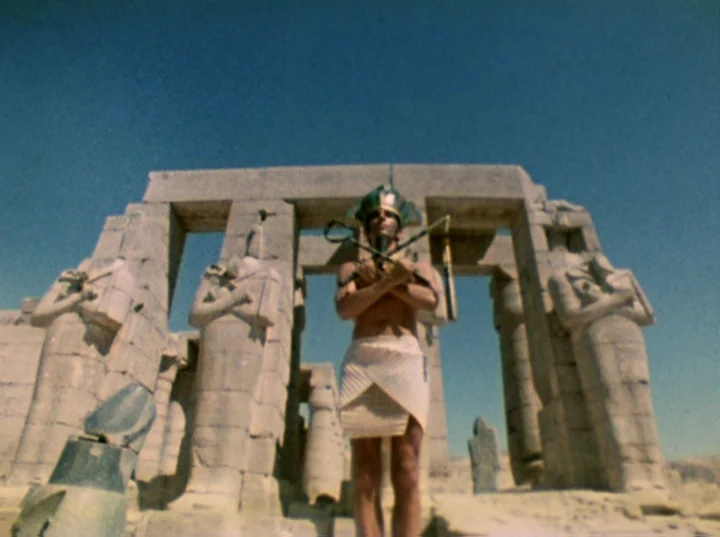 Lucifer Rising (1972) movie still: fullbody — An ancient stone temple ruin stands under a clear, bright blue sky. A man dressed as an Egyptian…