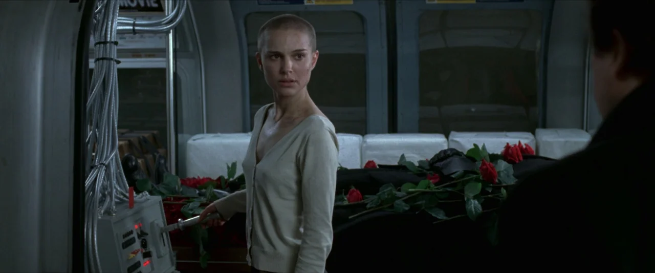V for Vendetta (2006) movie still: medium — A dark, industrial train car contains a control panel and a body covered by flowers. A young woman with…