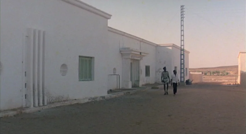 The Passenger (1975) movie still: wide — A stark white, low-slung building stands beside a dusty yard in a dry environment. Two men walk across the…
