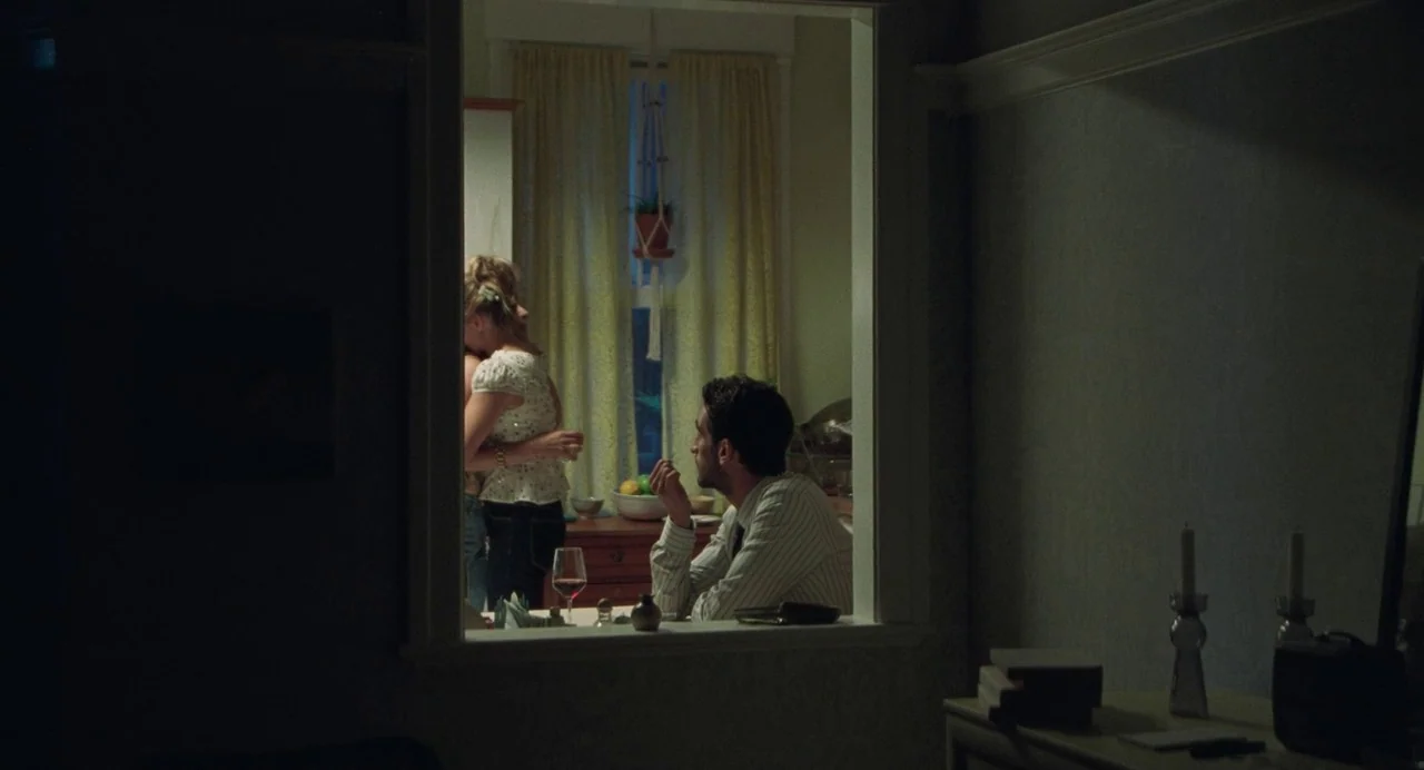 Matthias & Maxime (2019) movie still: medium — A dark residential interior view looking through a rectangular cutout into a dining area. A man sits…