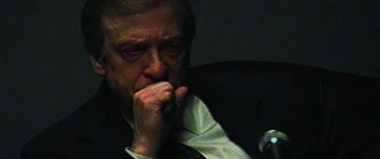 Dark Waters (2019) movie still: closeup — A dimly lit, dark space possibly at a media event or office interior. An elderly man in a dark suit sits…