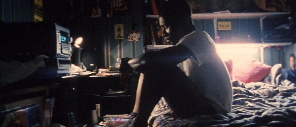 Wonderland (1999) movie still: medium — A dimly lit room with walls covered in posters and cluttered shelving units. A young man in a t-shirt sits…