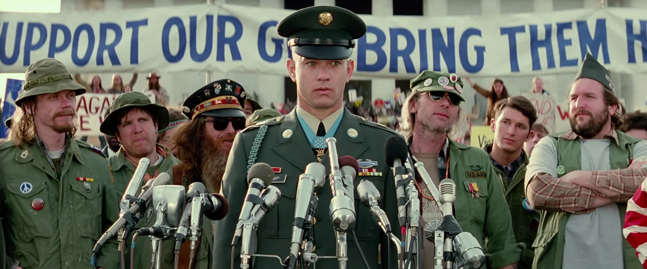Forrest Gump (1994) movie still: medium — A man in a military uniform stands at a podium before a large banner. Multiple microphones are arrayed in…