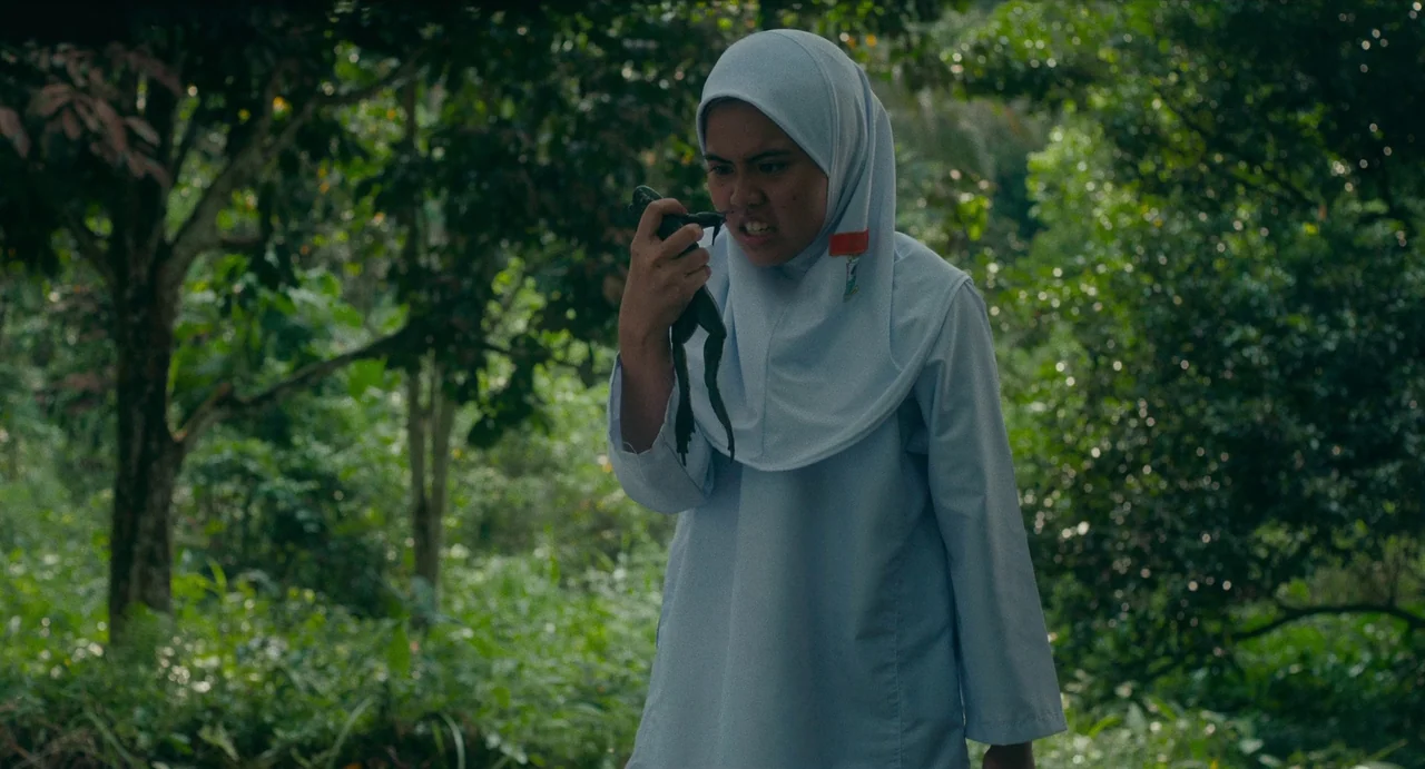 Tiger Stripes (2023) movie still: medium — A dense, lush tropical thicket with blurred trees filling the background space. A young woman in a…