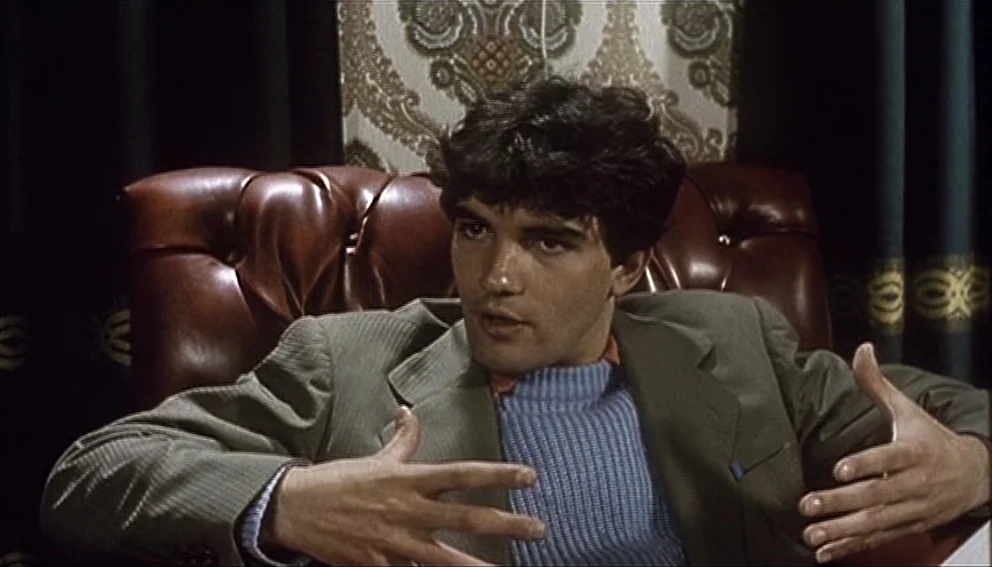 Labyrinth of Passion (1982) movie still: medium — A man sits in a dark leather chair against a patterned wallpaper background. He gestures with his…