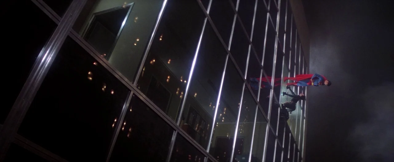 Superman (1978) movie still: wide — The reflective glass surface of a multistory office building dominates the dark frame. A superhero floats…