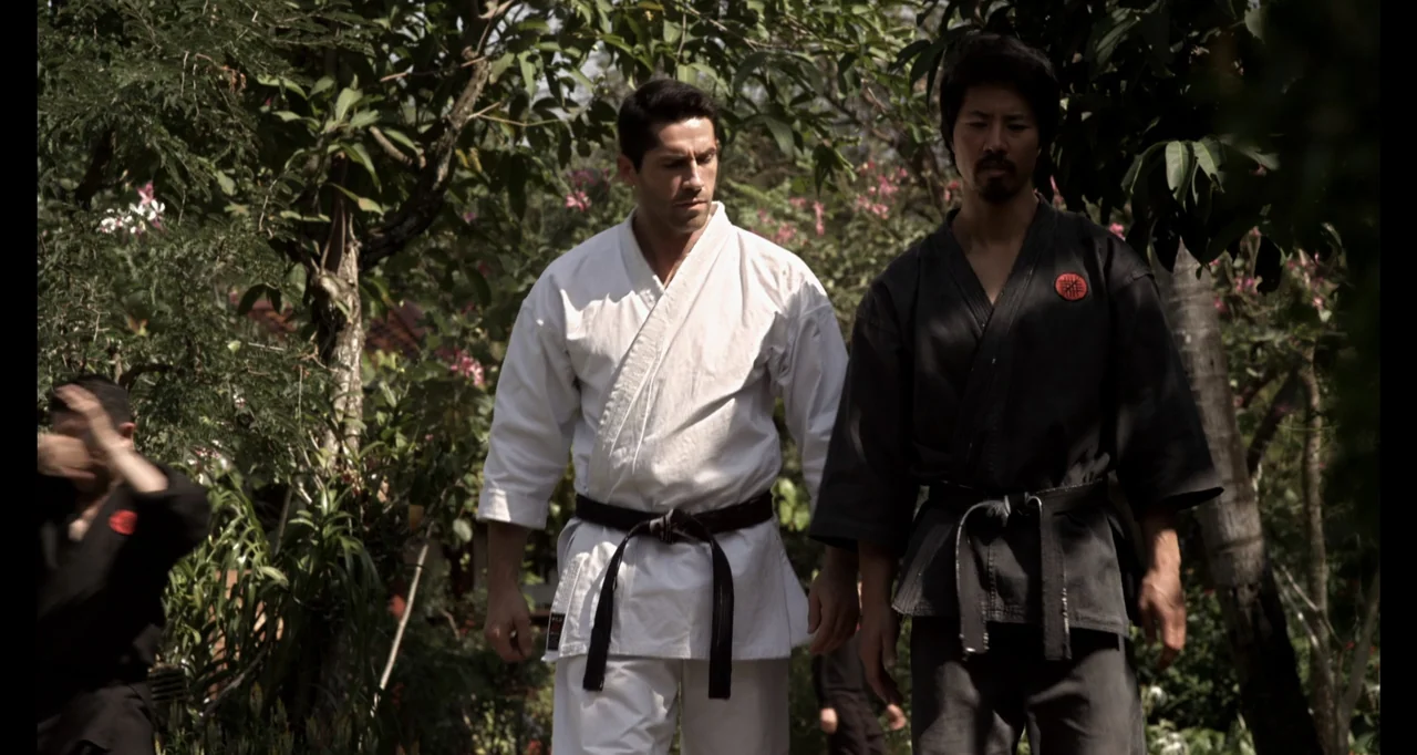 Ninja: Shadow of a Tear (2013) movie still: fullbody — A dense tropical garden with mature trees and foliage frames two men walking forward. The men…