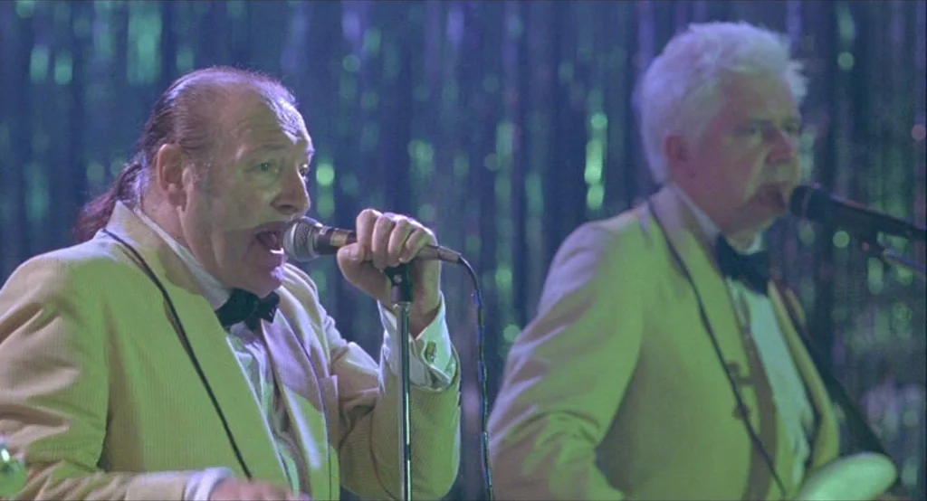 Looking for Eric (2009) movie still: medium — Two older male singers perform on a stage against a shimmering metallic curtain backdrop. The…