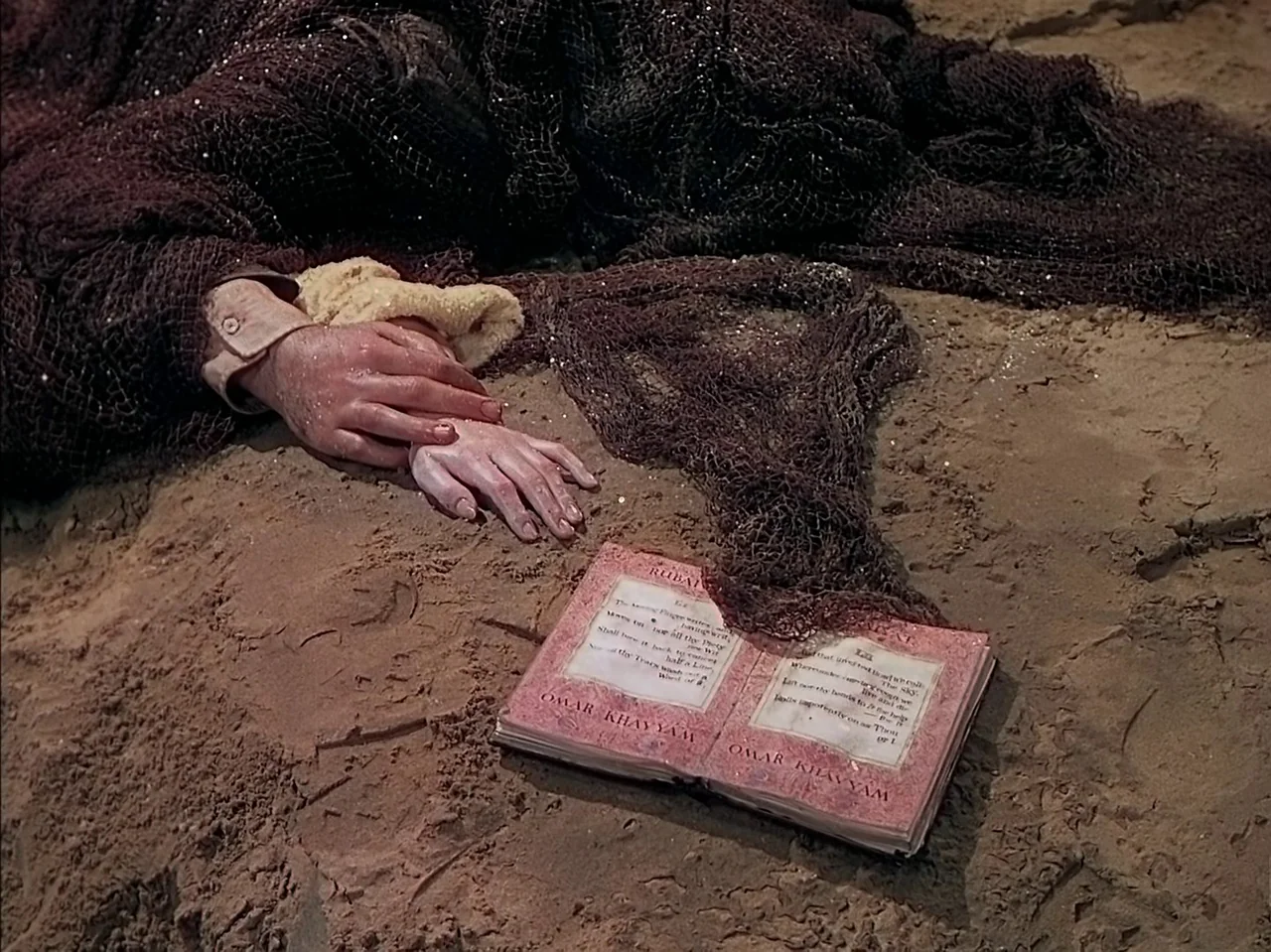 Pandora and the Flying Dutchman (1951) movie still: closeup — A dark fishing net and a pair of hands rest on sandy ground. An open book with printed…