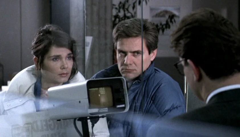 The Seventh Continent (1989) movie still: medium — A man and a woman look intently at a security monitor through a glass partition. The setting is a…