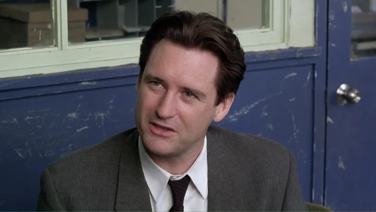 Mistrial (1996) movie still: medium — An office interior features a worn blue painted wall with a window pane behind. A middle-aged man in a suit…