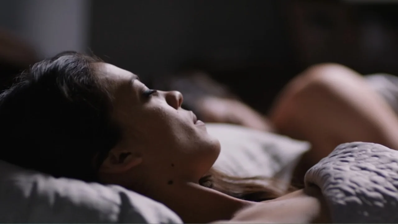 Alice in Marialand (2014) movie still: closeup — A dimly lit indoor bedroom features a woman resting on white pillows and sheets. The subject lies…
