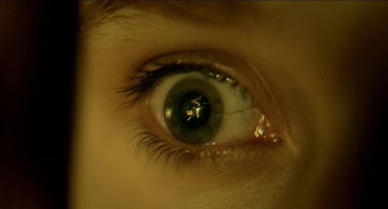 Trim Season (2023) movie still: closeup — A close-up view of a human eye with a greenish-brown iris and eyelashes. Light reflects off the pupil,…