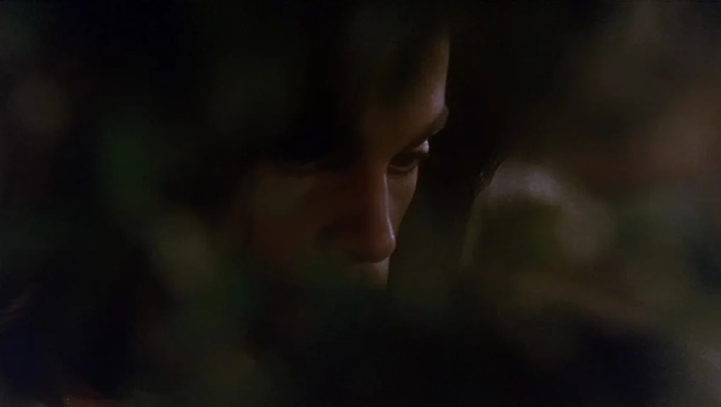 Graffiti Bridge (1990) movie still: closeup — A close view shows a face partially hidden behind dark, out-of-focus foreground leaves. The subject…