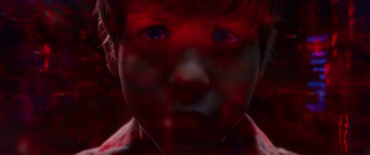 The Predator (2018) movie still: closeup — A close-up of a young boy's face obscured by a digital red light projection. Red light patterns wash over…