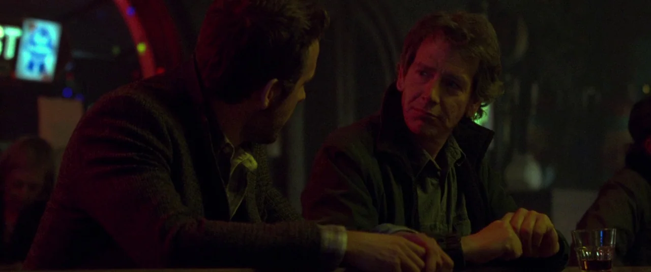 Mississippi Grind (2015) movie still: medium — A dimly lit bar at night with red neon illumination and cluttered background details. Two men sit…