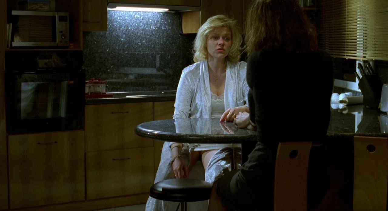 Read My Lips (2001) movie still: medium — A dimly lit room with wood-veneer cabinets and a dark granite breakfast bar counter. One woman in a white…
