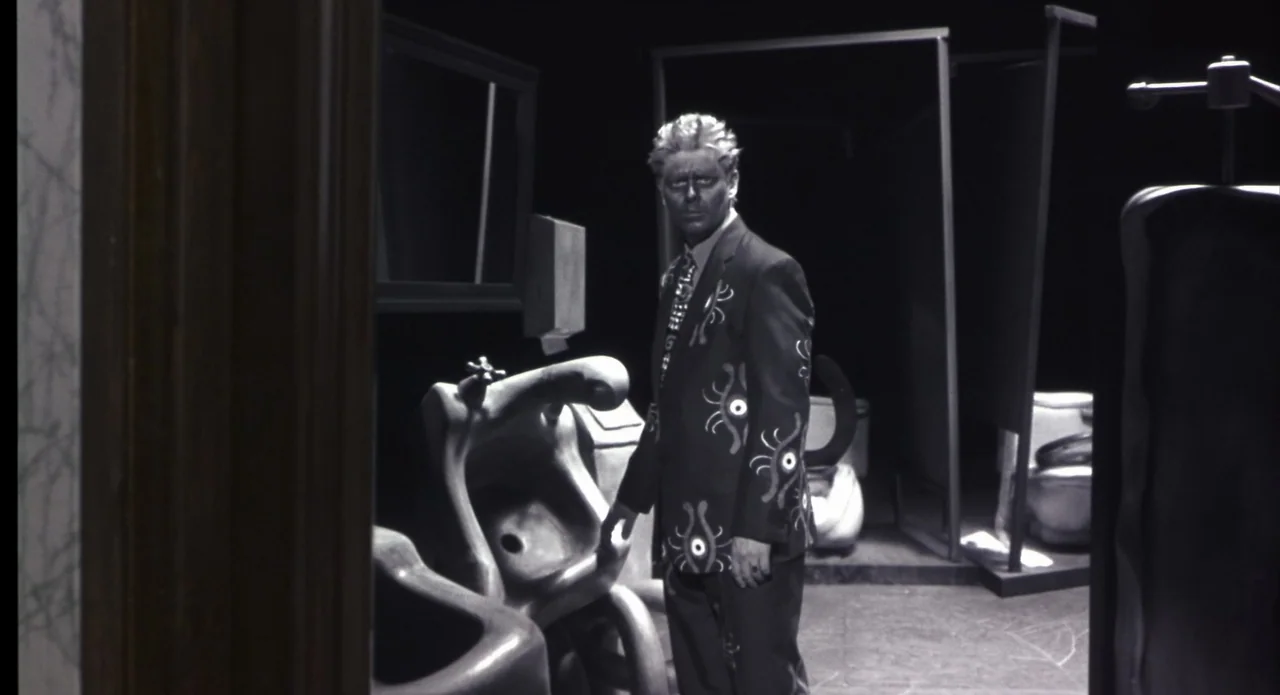Monkeybone (2001) movie still: medium — A dark, stylized restroom features surreal, organic-shaped fixtures and high-contrast lighting. A man with…
