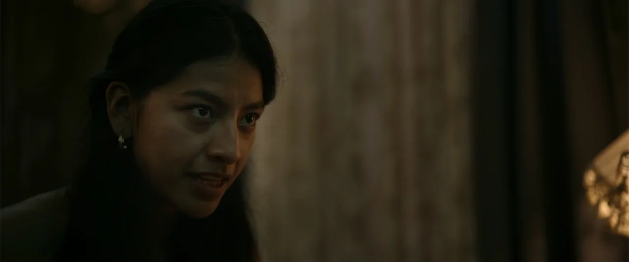 Evil Eye (2020) movie still: closeup — A dimly lit indoor space with textured walls visible behind the subject. A young woman with dark hair turns…