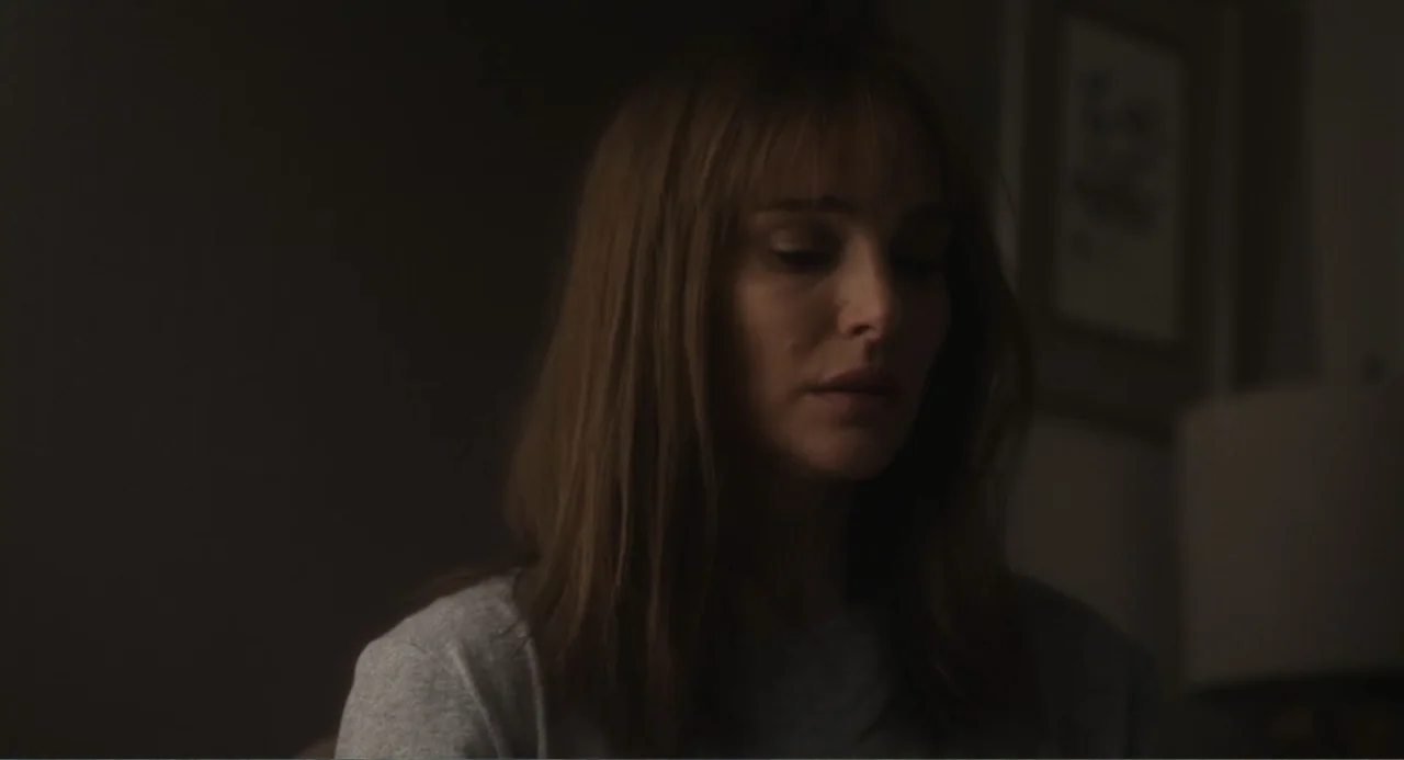 May December (2023) movie still: medium — A dimly lit room with muted colors and a dark wall. A woman looks downward while wearing a casual sweater.