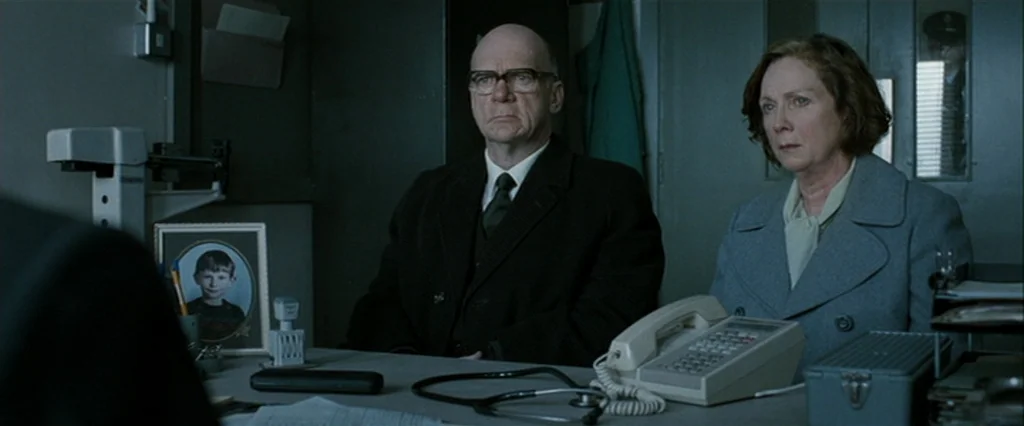 Hunger (2008) movie still: medium — An office with grey walls and a desk cluttered with medical and administrative items. A man and a woman sit…