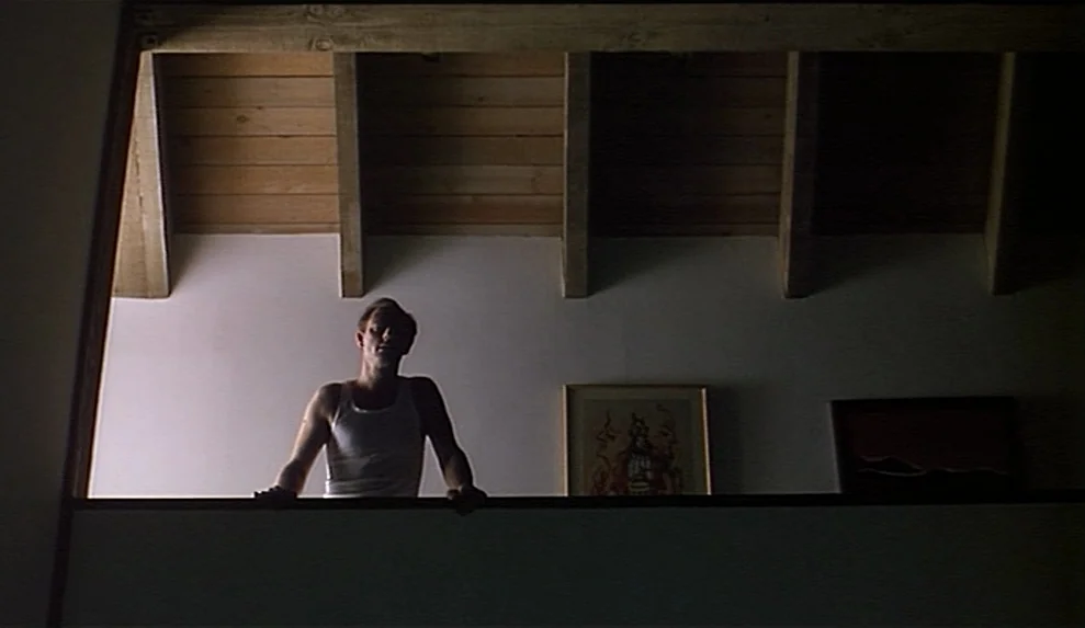 Safe movie still: medium — A man stands on an upper level looking over a wooden railing towards below. The wall behind him has wooden ceiling beams…