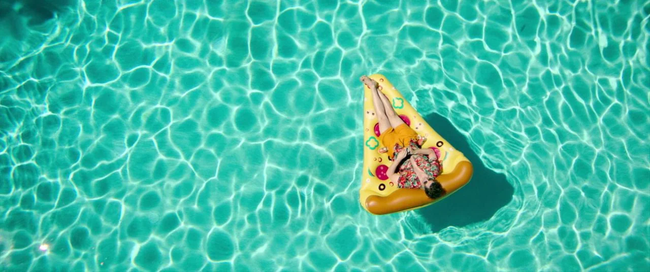 Palm Springs (2020) movie still: wide — A turquoise swimming pool filled with shimmering light patterns covers the entire frame. A lone man reclines…