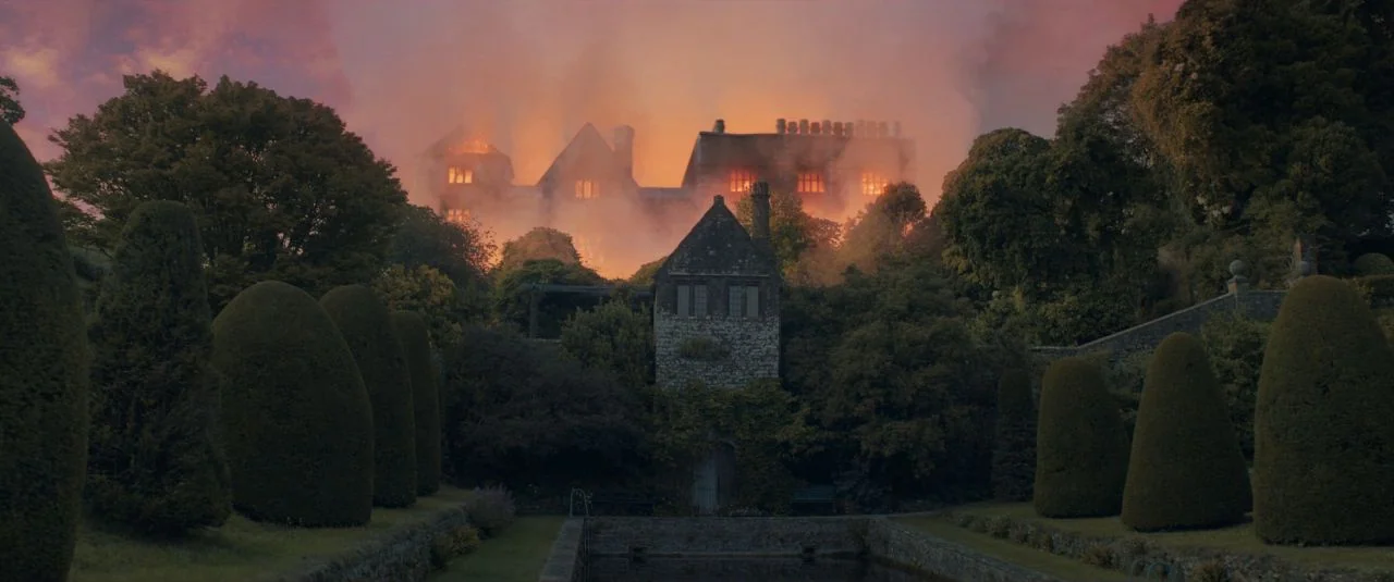 Rebecca movie still: establishing — A stone manor sits atop a hill, partially obscured by dense orange smoke and fire. A manicured garden with a…