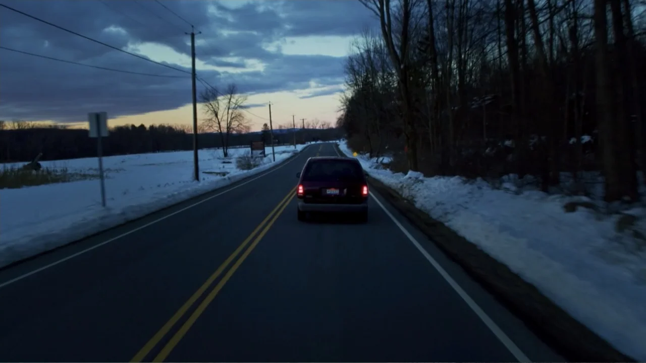 Burn (2019) movie still: wide — A rural asphalt road cuts through a snow-covered landscape during the blue hour. A dark minivan travels away from…