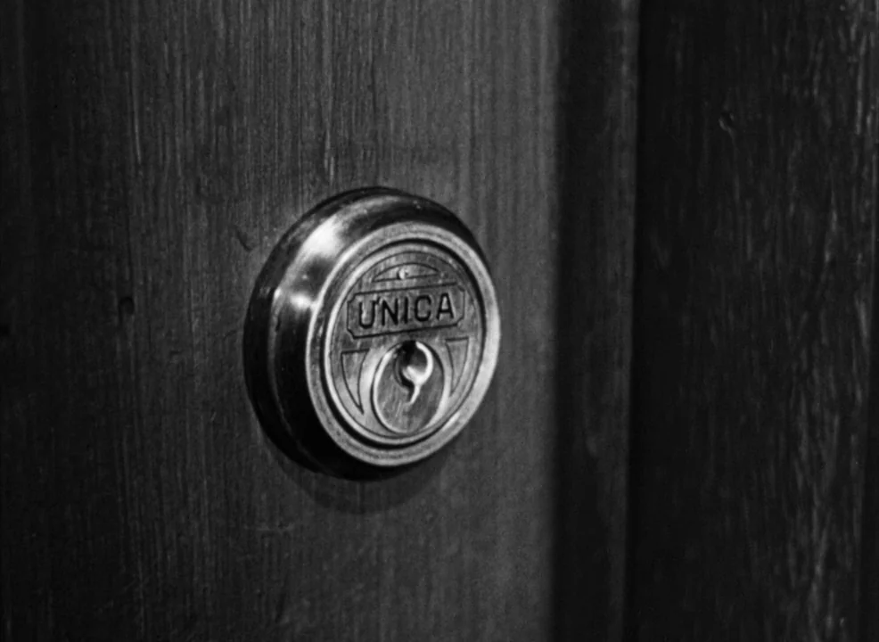 Notorious (1946) movie still: closeup — A close-up view of a metal circular lock embedded in a dark wooden door. The brand name UNICA is embossed on…