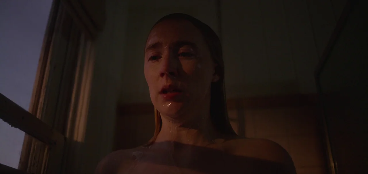 Foe (2023) movie still: closeup — A dark bathroom interior with a shower stall illuminated by a nearby window. A woman with wet hair stands with her…
