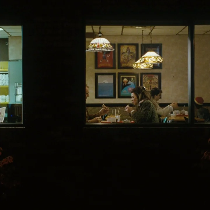 Mommy (2014) movie still: wide — A view through a window reveals people dining at tables inside a restaurant. The interior is decorated with framed…