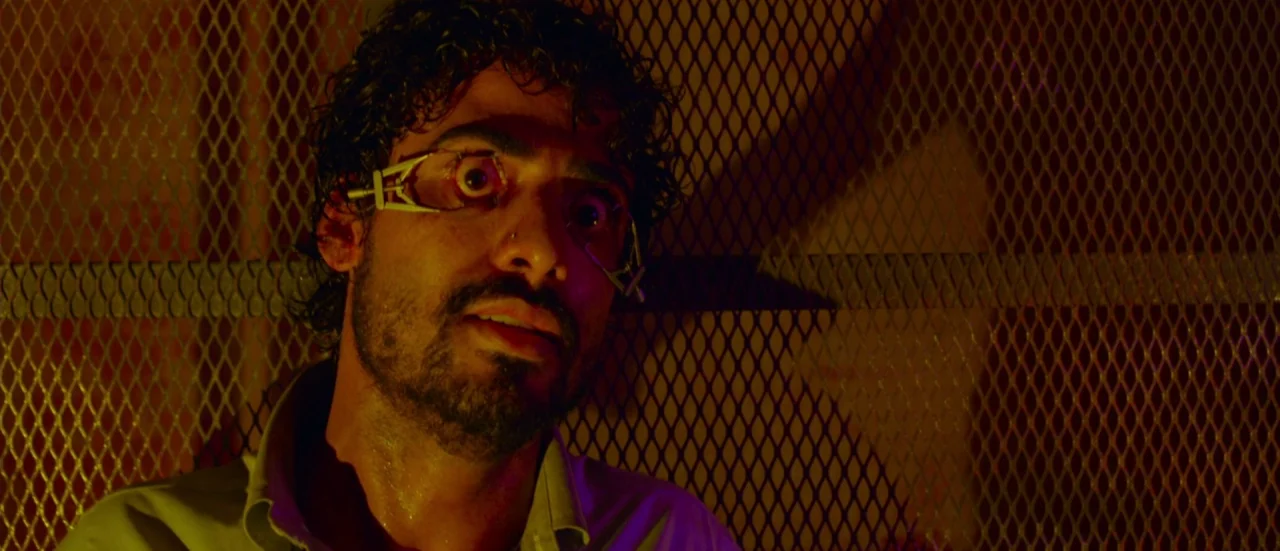 Rupture (2016) movie still: closeup — A man stands before a dark industrial metal mesh wall in a dim room. He has a metal speculum device forcing…