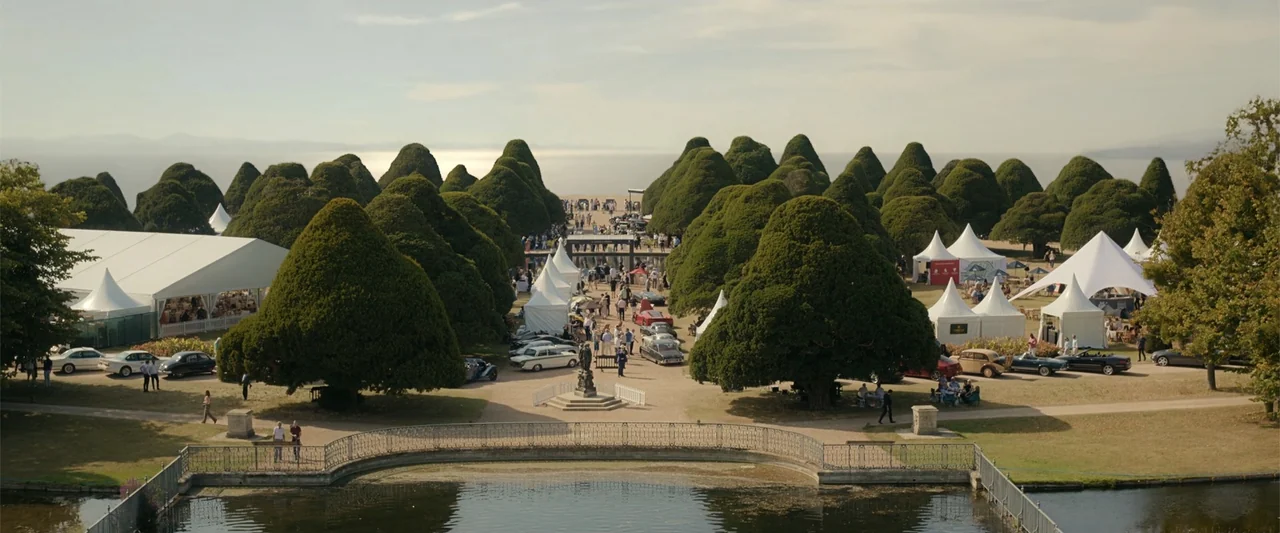 Maintenance Required (2025) movie still: wide — A sprawling outdoor estate park features manicured topiary trees and multiple white event tents. A…