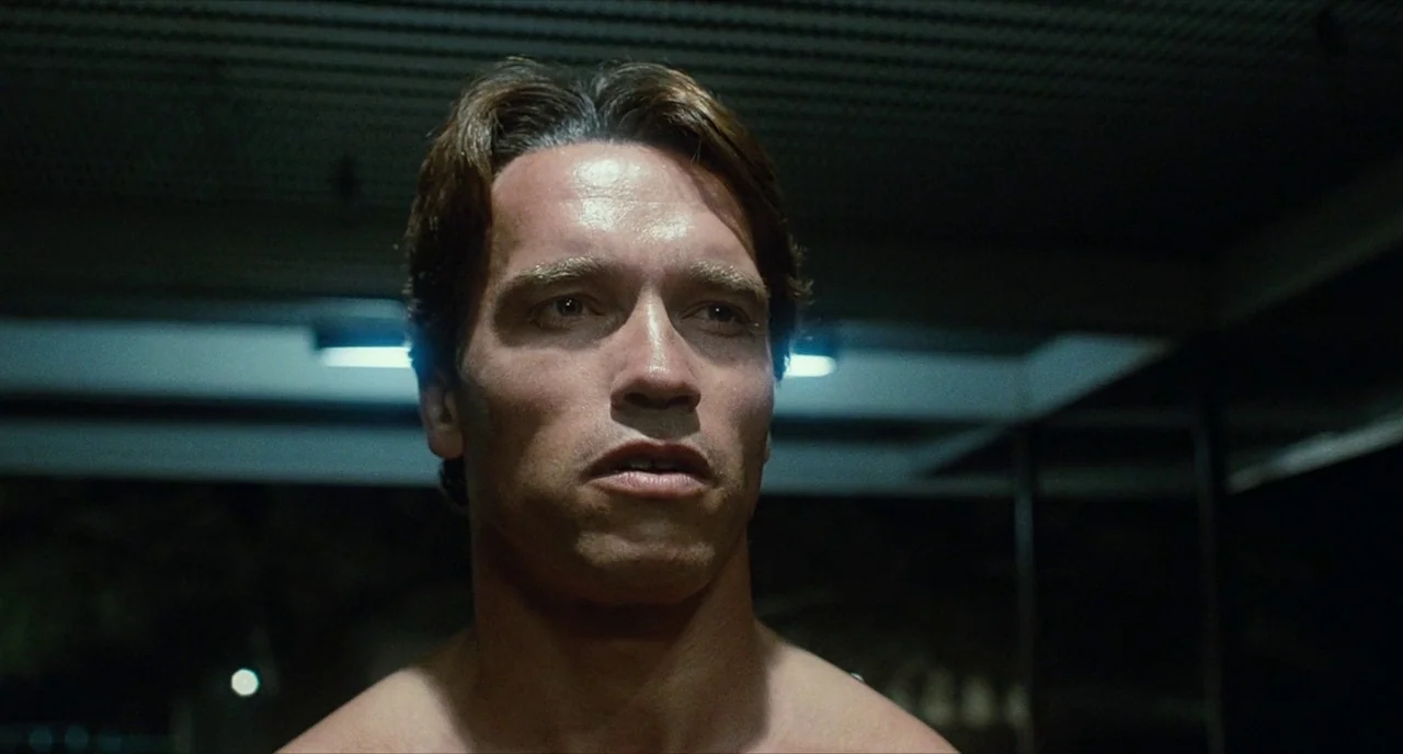 The Terminator (1984) movie still: closeup — A dark night setting at an outdoor location with overhead structural metal ceiling beams. A shirtless…