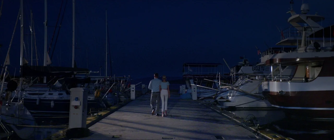 Holiday movie still: fullbody — A quiet marina at night features a long concrete pier lined with moored yachts. A man and a woman walk away from the…