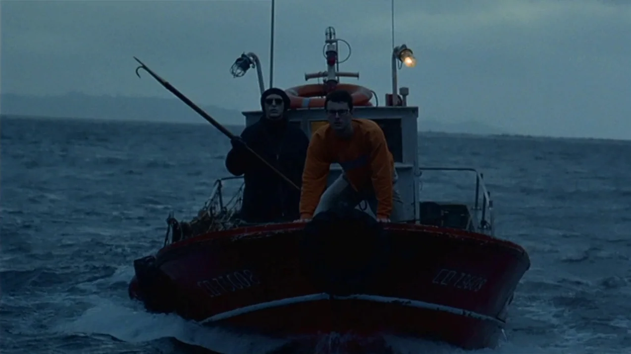 Dagon (2001) movie still: fullbody — A small red boat cuts through choppy dark water under a dim twilight sky. Two men stand on the deck looking…