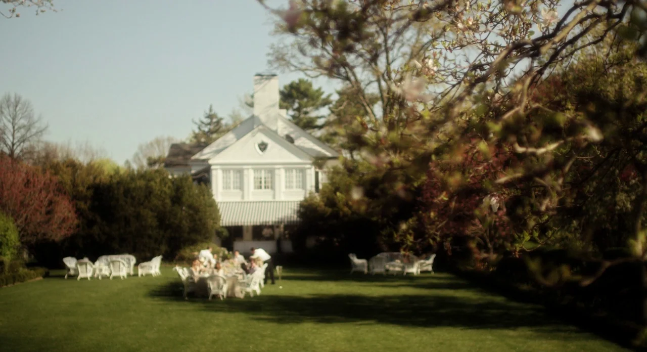 Side Effects (2013) movie still: wide — A sprawling green lawn stretches toward a white colonial house with an awning. A small group of people sits…