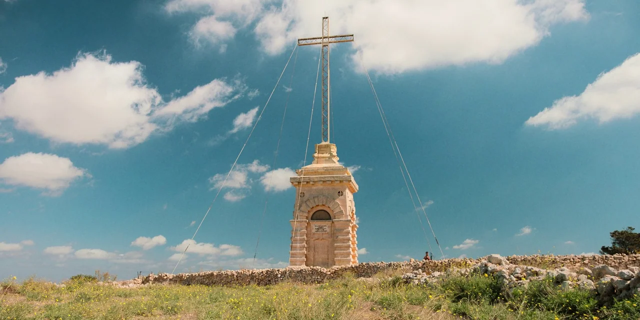 Sniper: G.R.I.T. (2023) movie still: establishing — A stone monument topped with a metal cross stands on a barren grassy hillside. The structure is…