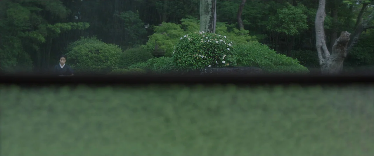 The Handmaiden (2016) movie still: wide — A rain-drenched Japanese-style courtyard featuring manicured shrubs and trees in the background. A woman…