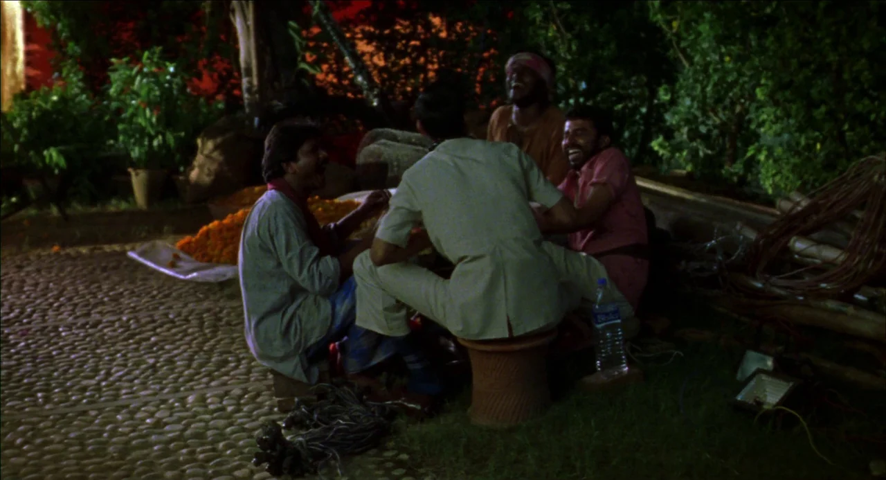 Monsoon Wedding (2001) movie still: medium — A stone-paved outdoor space is surrounded by thick green foliage at night. Four men sit together on…