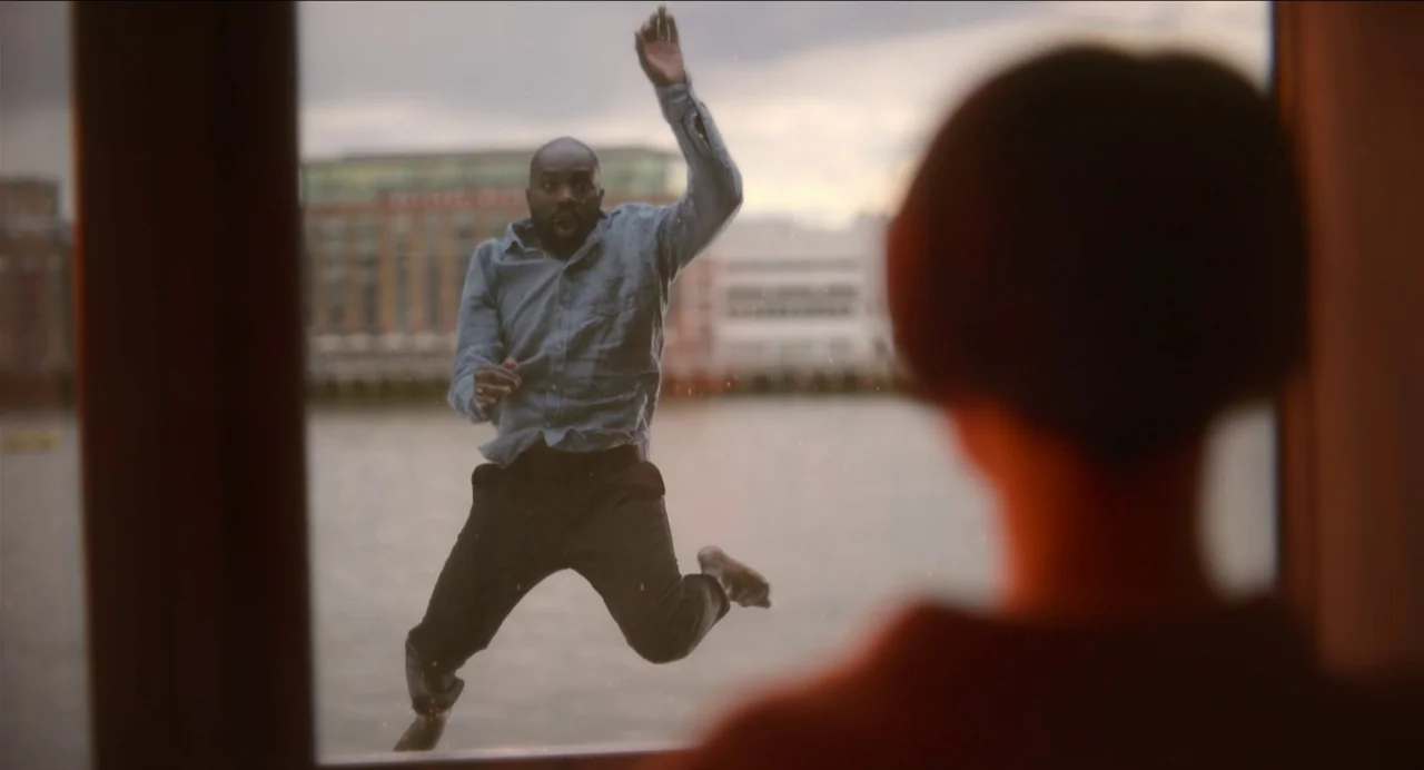 Men (2022) movie still: medium — A man suspended in mid-air appears outside a glass window pane. A foreground figure views the man through the…