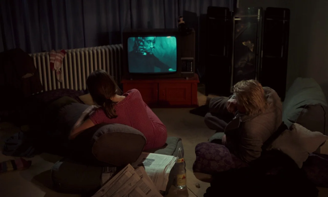 Wrong Move (1975) movie still: wide — A dimly lit room at night features a television set displaying a black-and-white film. Two people sit on floor…
