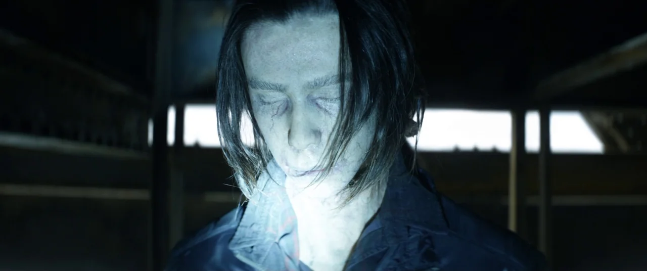 The Empty Man (2020) movie still: closeup — A dark indoor setting features the man positioned against a blurred background with horizontal beams. He…