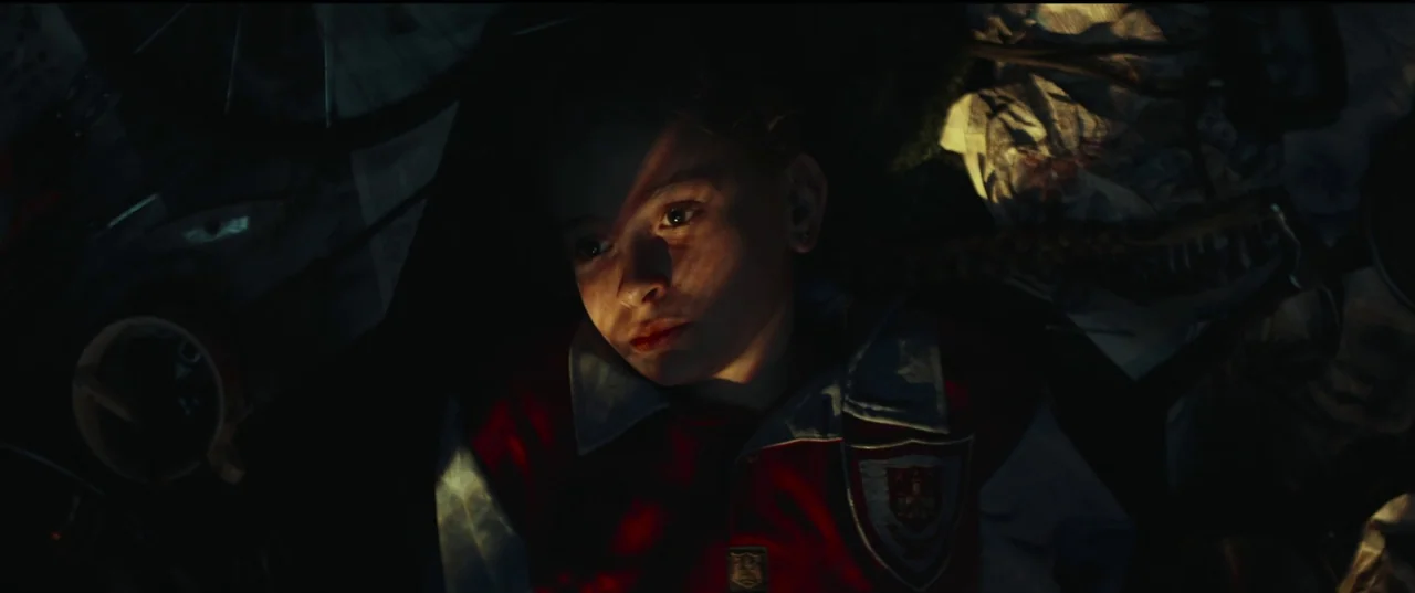 Scrapper (2023) movie still: closeup — A dark, cluttered interior space is filled with indistinct, shadowed objects and equipment. A young girl in a…