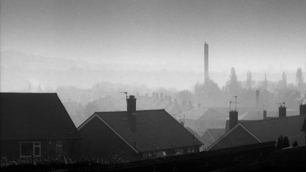 TwentyFourSeven (1997) movie still: establishing — An expanse of suburban residential rooftops stretches toward a distant industrial brick chimney.…