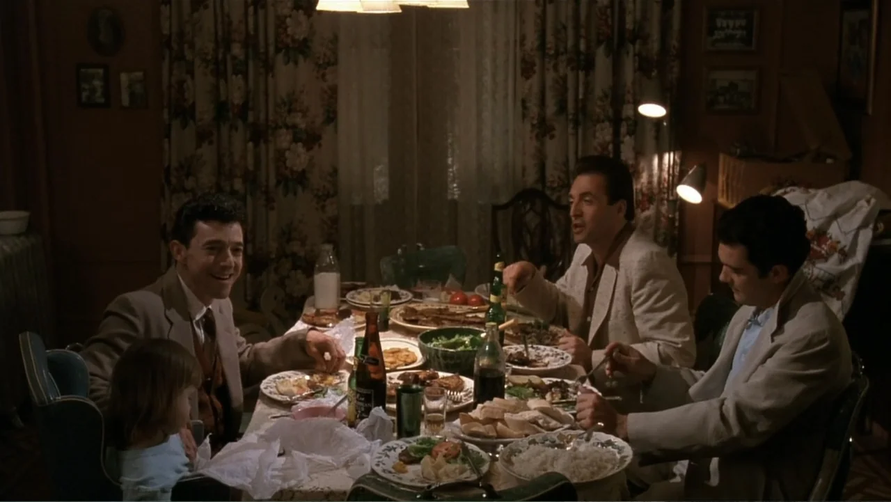 The Mambo Kings (1992) movie still: medium — A 1950s dining room features floral wallpaper, a draped curtain, and a crowded table. Three men and a…