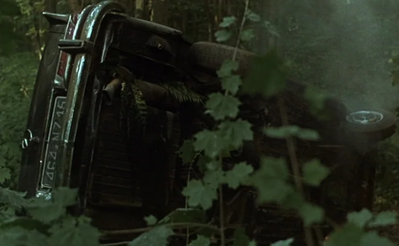 Maîtresse (1975) movie still: medium — A vintage dark car rests upside down amidst dense foliage in a wooded area. Light smoke emanates from the…