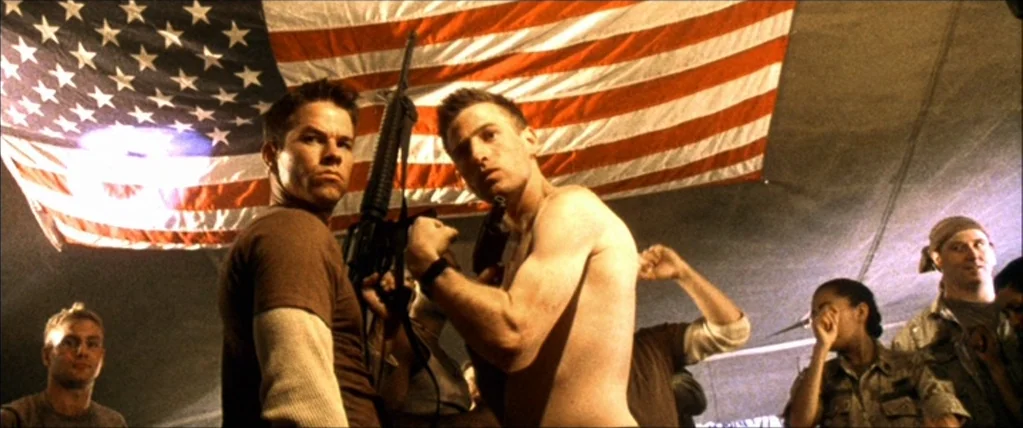 Three Kings (1999) movie still: medium — A military tent interior features a large American flag hung as a backdrop. Two soldiers pose with a rifle…